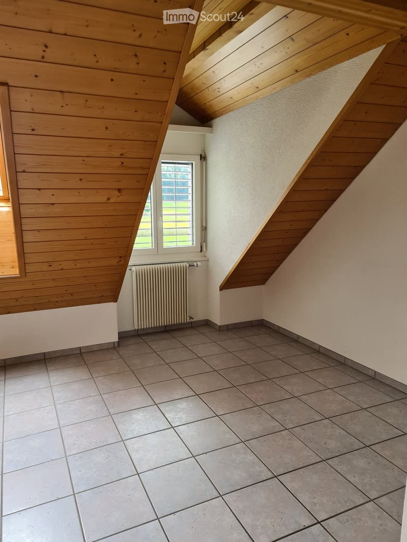 Empty attic room with wooden slanted ceilings, tiled floor, and a single window with white frame. A radiator is mounted on the wall below the window.