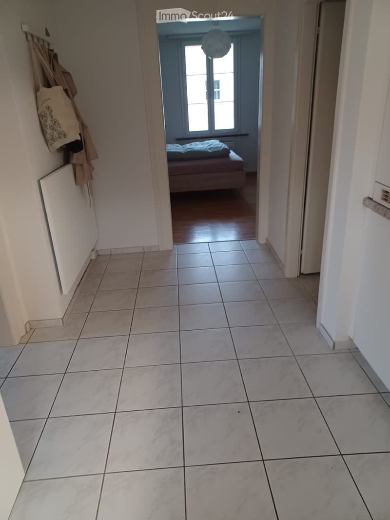 A clean, tiled hallway with white walls, leading to a room with a bed and a window. Hooks on the wall hold coats and bags.