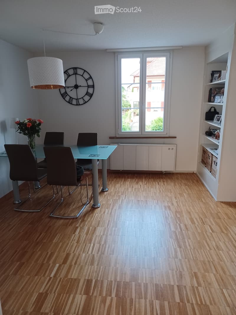 Dining room with wooden flooring, a glass dining table with four chairs, a large wall clock, a vase with flowers, and built-in cabinets.