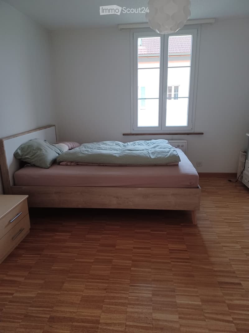 A small, well-lit bedroom with wooden flooring, a single bed with a light green duvet and pillows, a wooden nightstand, and a window with white frames.
