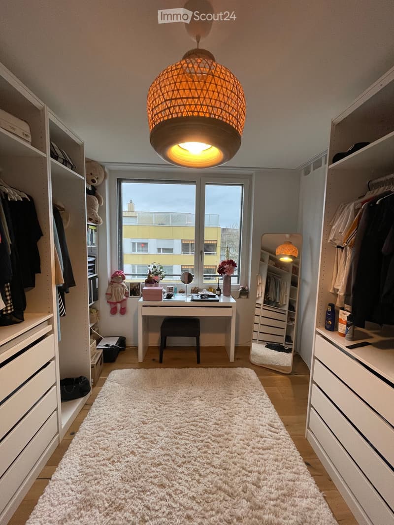 A well-lit room with a large window, a white shaggy rug, a modern hanging lamp, a desk with a chair, a wardrobe, and a mirror. The room features built-in storage and a cozy, organized layout.