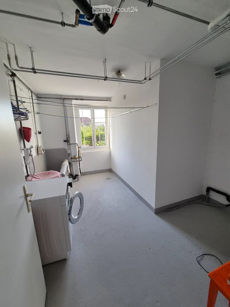 Empty room with exposed pipes, a window, a washing machine, and a clothes rack with a few items.