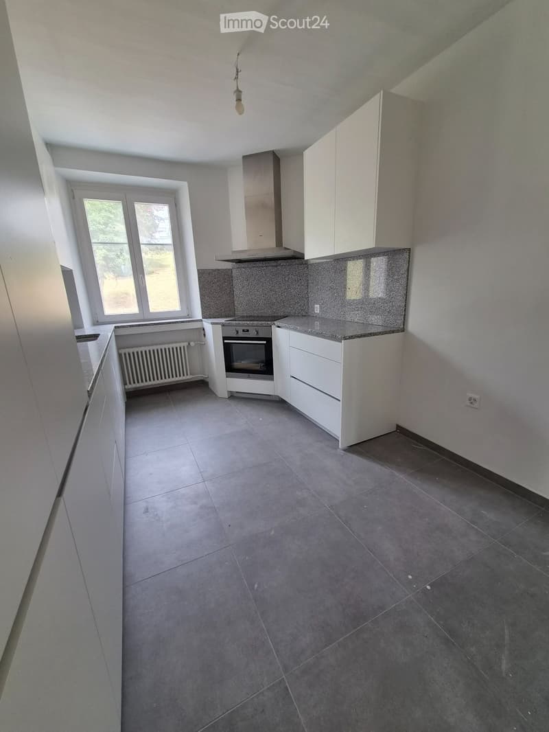Empty kitchen with white cabinets, granite countertop, built-in oven, and a window overlooking a green area.