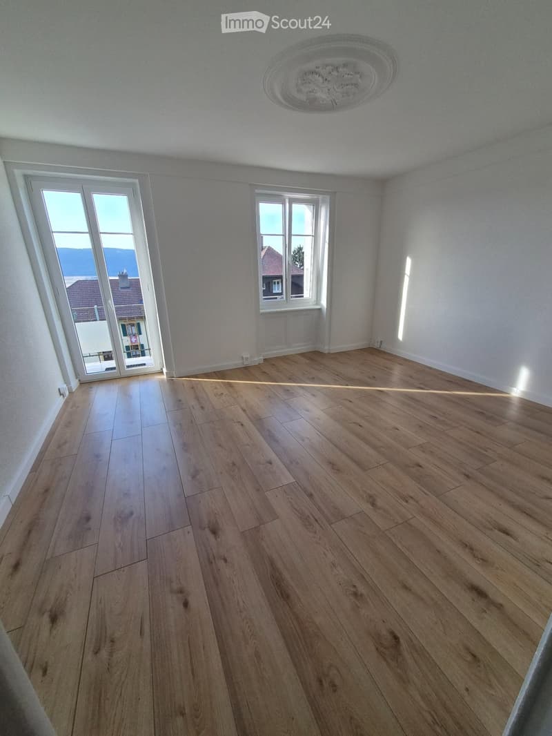 Empty room with light wooden flooring, white walls, and large windows allowing natural light to enter.