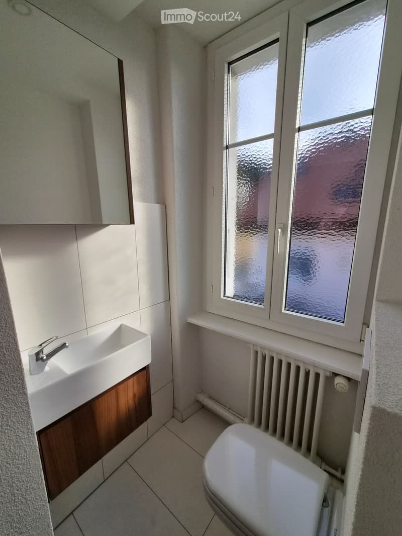 Modern bathroom with white tiled walls and floor, a white sink with a chrome faucet, a wooden cabinet, a large mirror, a toilet, and a radiator below the window.