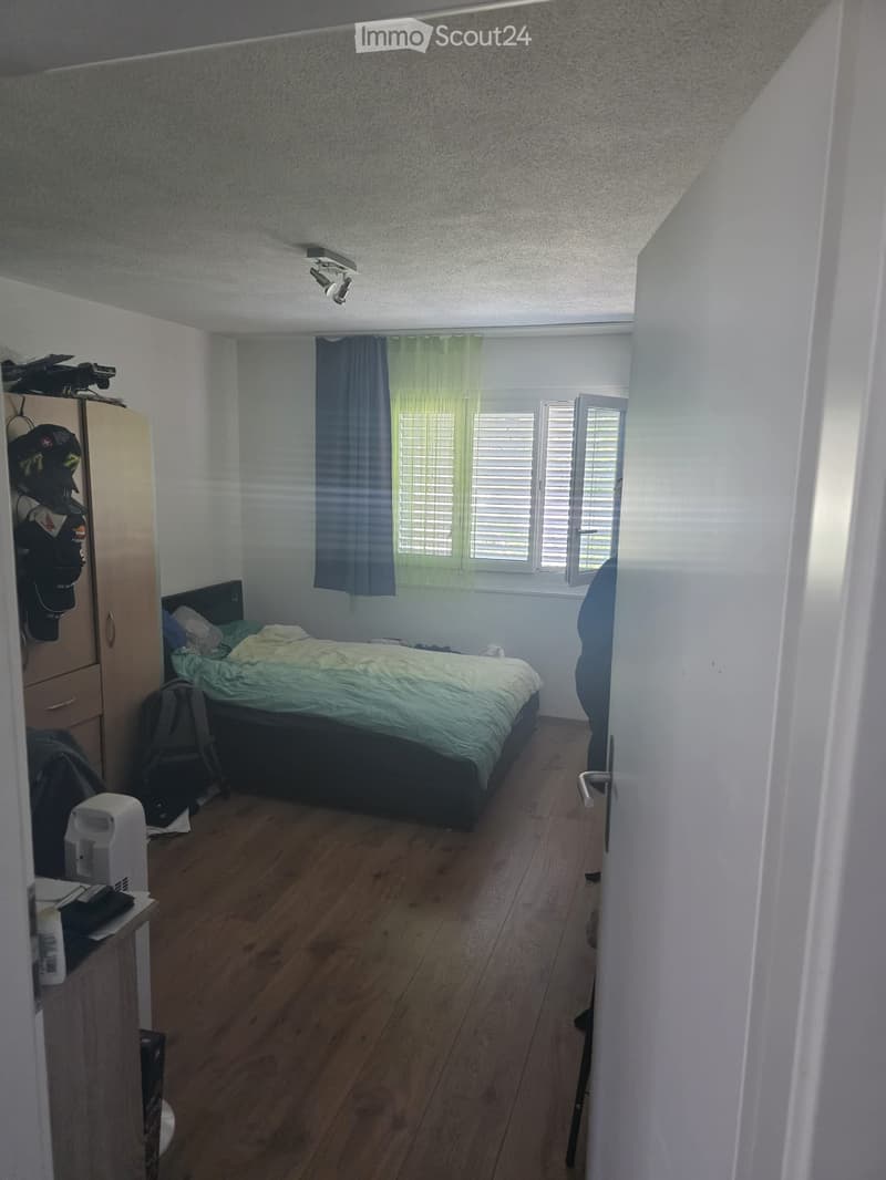 A small bedroom with a bed, wardrobe, and a backpack on the floor. The room features wooden flooring and white walls, with a window covered by curtains.