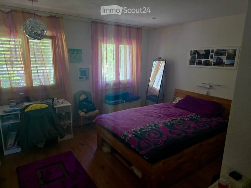 Cozy bedroom with wooden flooring, a large bed with purple bedding, a nightstand, a chair, and a mirror. The room features sheer curtains and several picture frames on the walls.