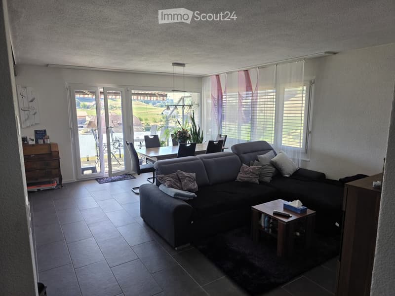 Modern living room with a grey sectional sofa, coffee table, dining area with chairs, side table, and a view of the outdoor area through large windows and sliding doors.