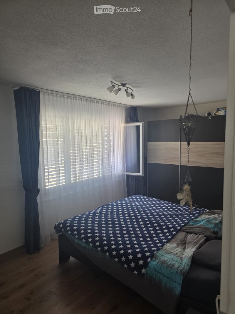 Bedroom with a blue and white star-patterned bedspread, wooden floor, dark blue and grey walls, large window with white curtains, and a hanging decorative item.