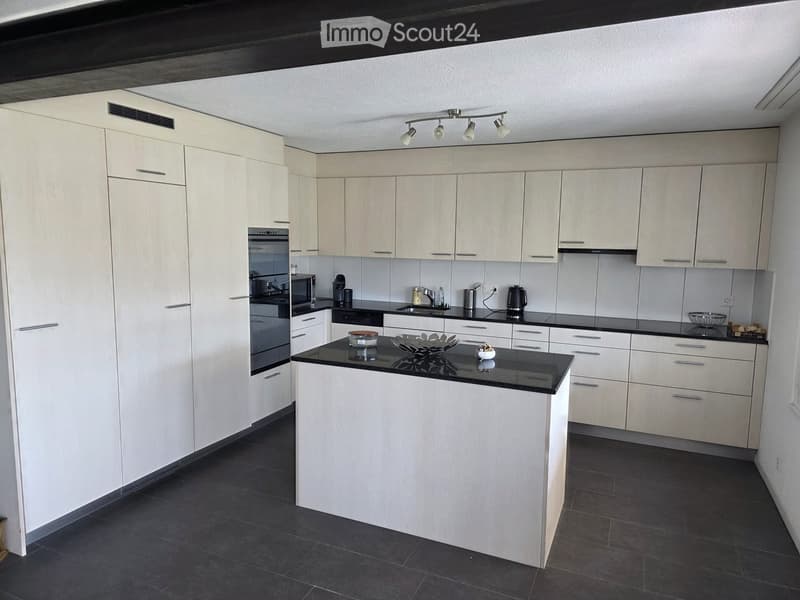 Modern kitchen with white cabinets, black countertops, and a central island. Appliances include an oven, microwave, and coffee maker. The space features sleek, contemporary design with integrated lighting and ample storage.