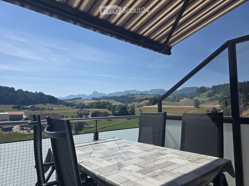 Balcony with outdoor furniture, overlooking a scenic countryside with mountains and fields.