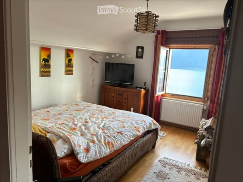 Cozy bedroom with wooden flooring, a large window offering a scenic view, a bed with a colorful quilt, a wooden dresser with a TV, and a floor lamp.