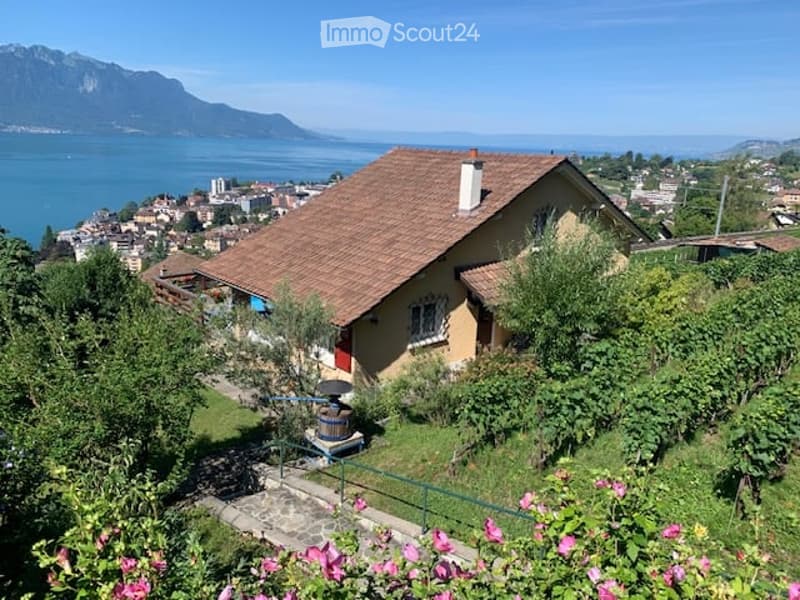 A charming yellow house with a red-tiled roof, nestled in a lush garden with vibrant pink flowers and a scenic view of a lake and mountains in the background.