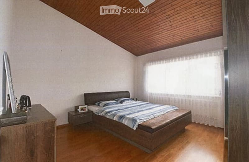 Cozy bedroom with wooden floors, slanted wooden ceiling, large window with white curtains, bed with striped bedding, nightstand, wooden dresser, and laptop on dresser.