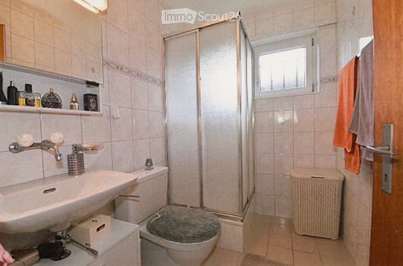 Bathroom with white tiled walls and floor, a sink with a faucet, a toilet with a grey seat cover, a glass-enclosed shower, a small shelf with toiletries, and a wicker basket on the floor.