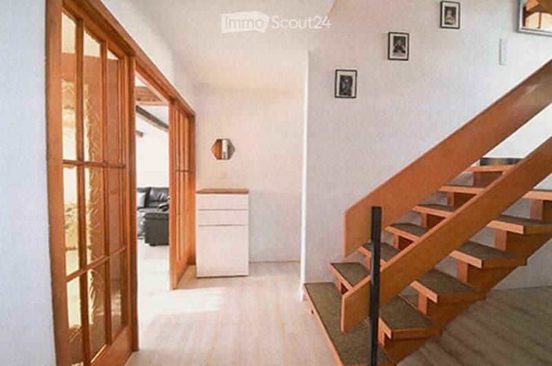 Bright hallway with wooden flooring, leading to a staircase and an open living area. Features include a small white cabinet, a black sofa, and framed pictures on the wall.