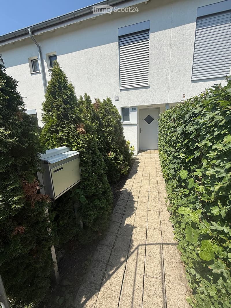 A pathway leads to a white building entrance, flanked by neatly trimmed hedges and a small utility box on the left.