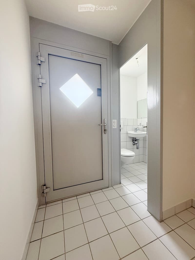 A narrow hallway with white tiled flooring, leading to an open door that reveals a small bathroom with a toilet, sink, and mirror.