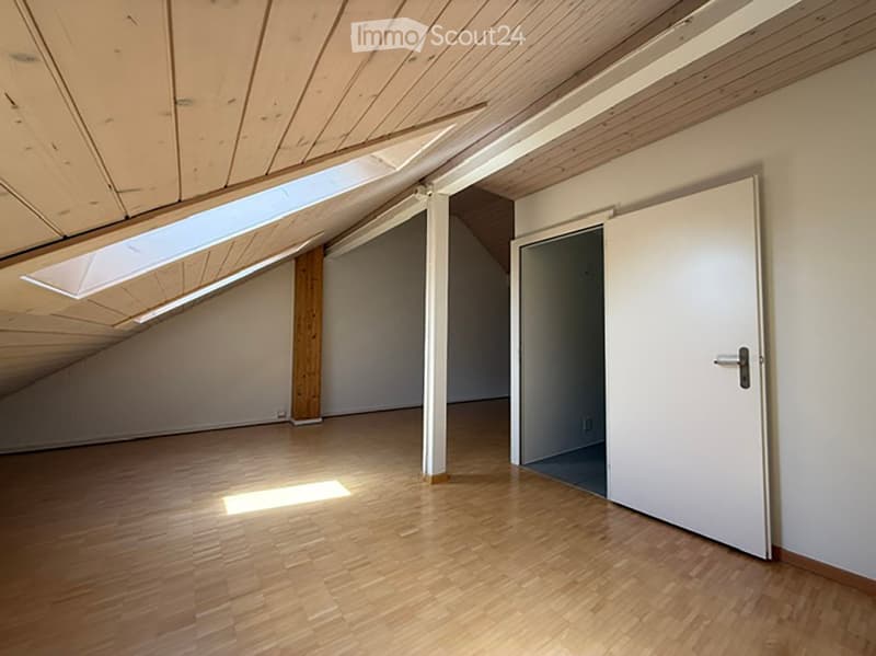 Empty attic space with wooden flooring, white walls, and exposed wooden beams. Features a skylight and a closed white door leading to another area.