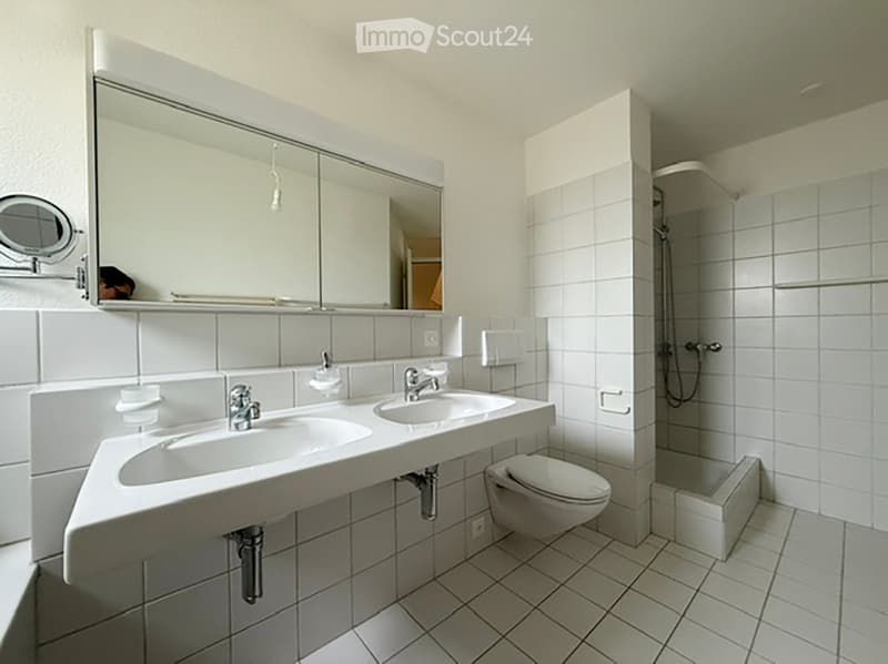 Modern bathroom with white tiled walls and floor, double sink vanity, large mirror, toilet, and walk-in shower with glass enclosure.