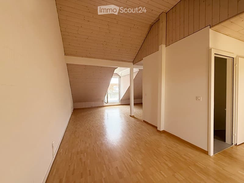 Empty room with wooden flooring and white walls, featuring a slanted wooden ceiling and a doorway leading to another space.