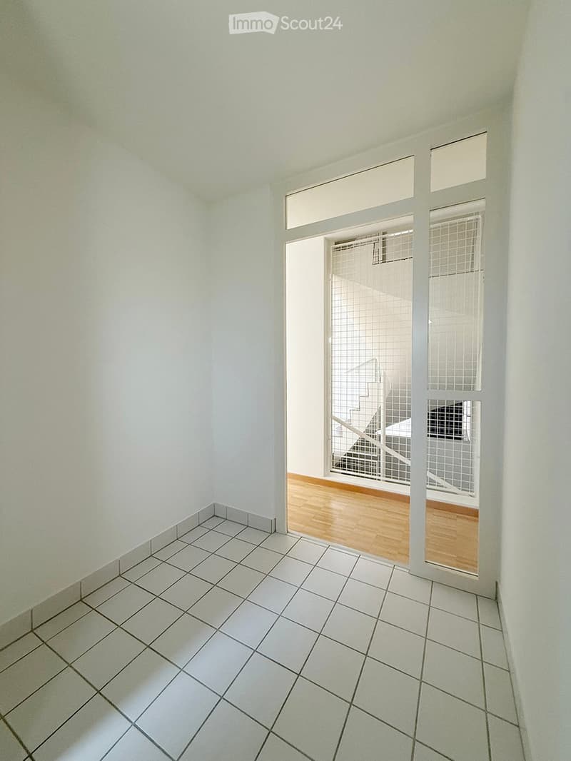 Empty room with white tiled floor and white walls, featuring a doorway leading to another room with wooden flooring and a visible staircase.