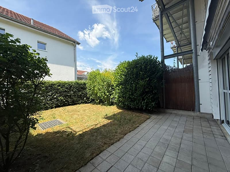 Garden area with a paved patio, grassy lawn, trimmed hedges, and a wooden gate leading to a modern house.