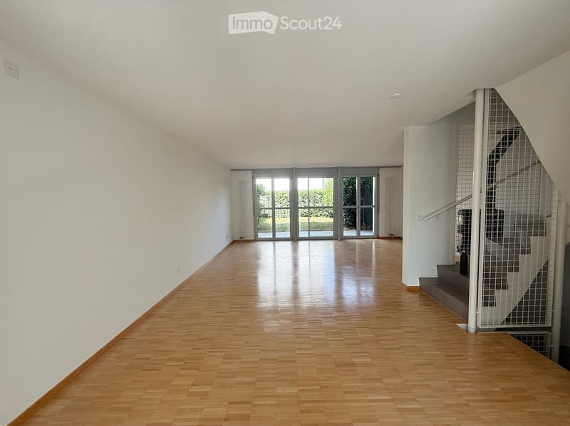 Bright, empty hallway with wooden flooring, white walls, large glass doors leading outside, and a staircase with a metal railing on the right side.