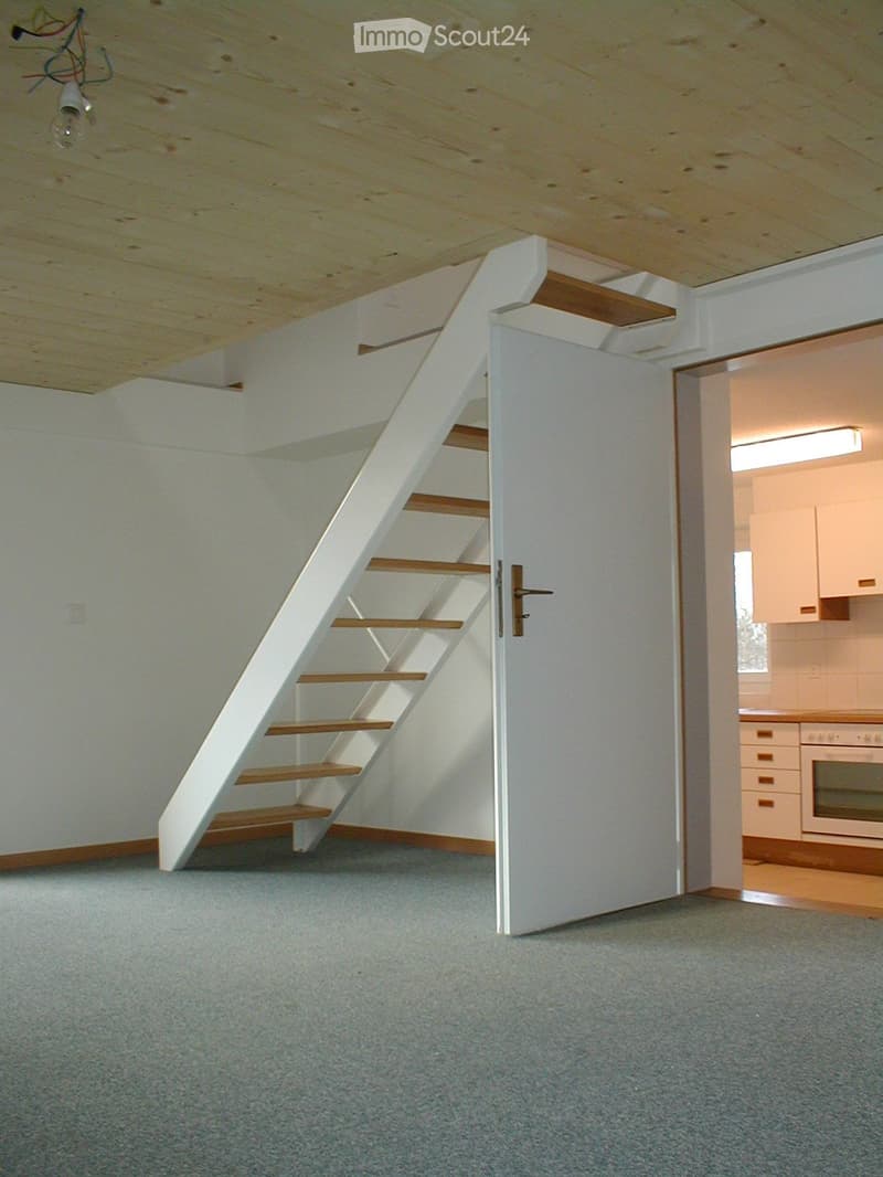 Open room with open staircase to upper floor, white walls, white door, gray flooring, ceiling light, kitchen in the background