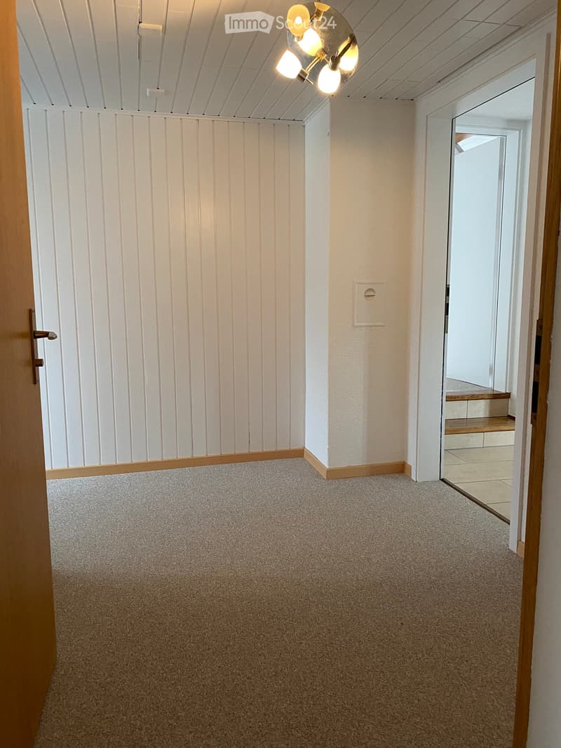 Empty hallway with carpeted floor, white paneled walls, and a ceiling light fixture. A doorway leads to a staircase area with wooden steps and a tiled landing.
