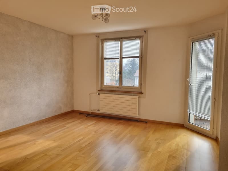 Empty room with wooden flooring, white walls, and large windows with blinds. A radiator is visible under one window, and a sliding door leads to an outdoor area.