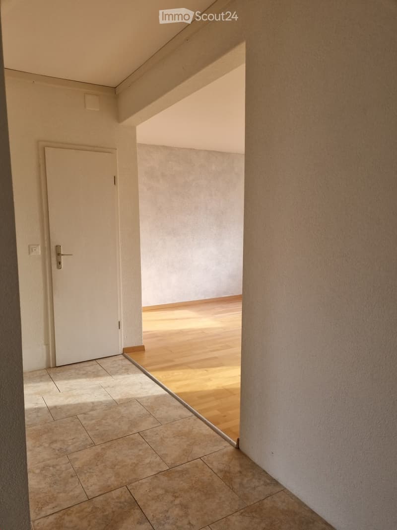 A narrow hallway with tiled flooring and a white door leading to a room with wooden flooring.