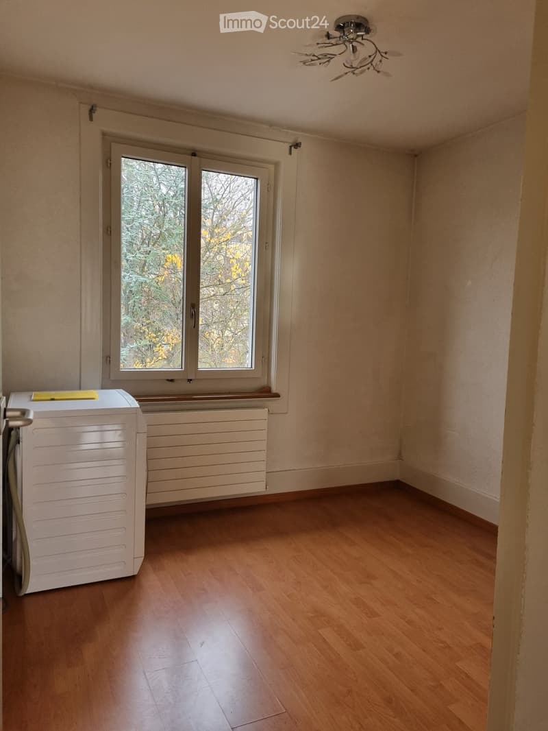 Empty room with wooden flooring, white walls, a window with a view of trees, a radiator, and a white appliance (likely a washing machine) in the corner.