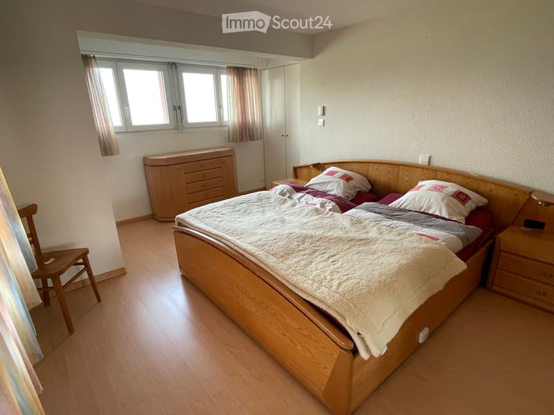 Cozy bedroom with wooden bed, nightstand, chair, wardrobe, and mirror. Light-colored bedding and curtains.