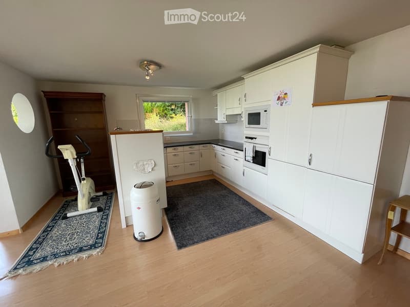 A well-lit kitchen with white cabinets, a microwave, an oven, an exercise bike, a small shelf, and a trash can. The floor is wooden, and there are two rugs in the space.