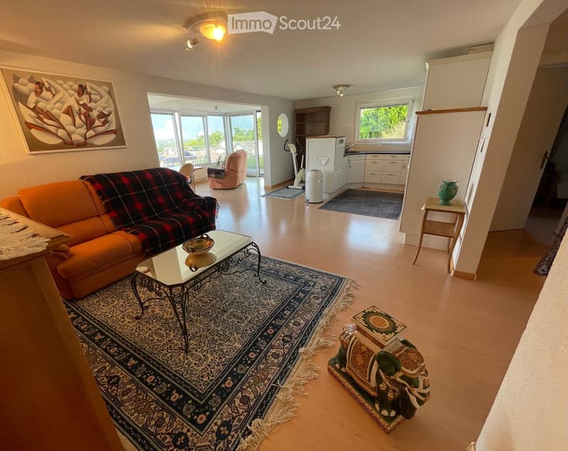 Bright living room with a view, featuring a sofa, coffee table, chair, and a small kitchen area with cabinets and a refrigerator.