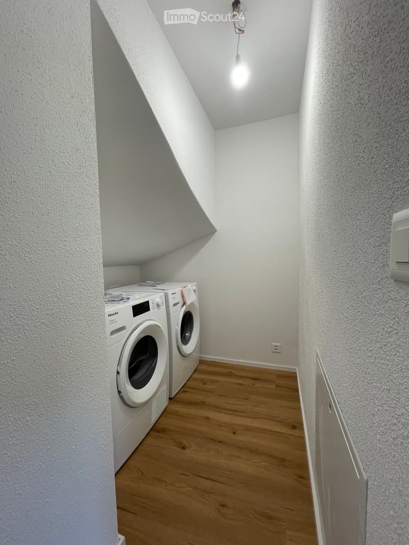 Small, well-lit room with wooden flooring, white walls, and a washing machine and dryer unit.