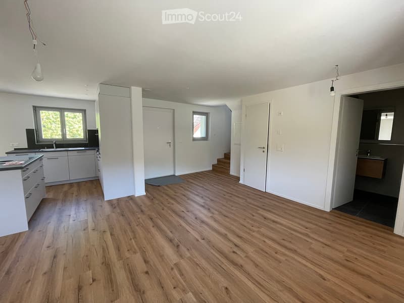 Empty room with wooden flooring, white walls, and a small kitchen area visible in the background. No furniture or personal items present.