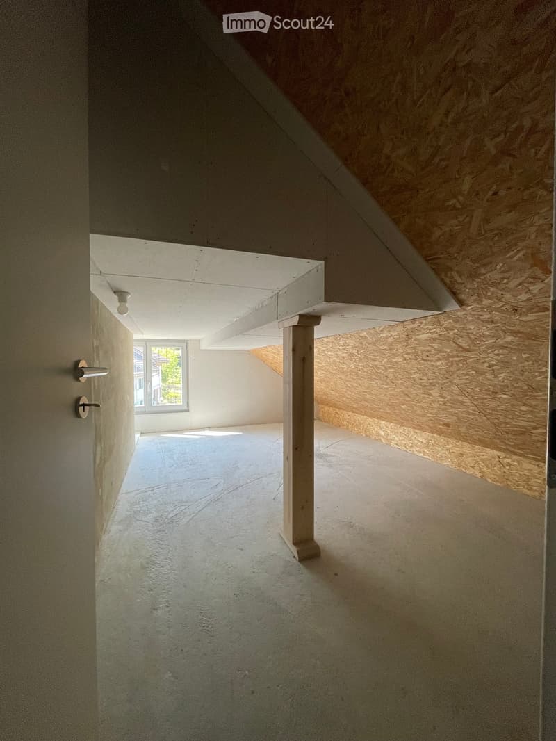 Empty room with concrete floor, white walls, exposed wooden ceiling beams, and a single window letting in natural light.