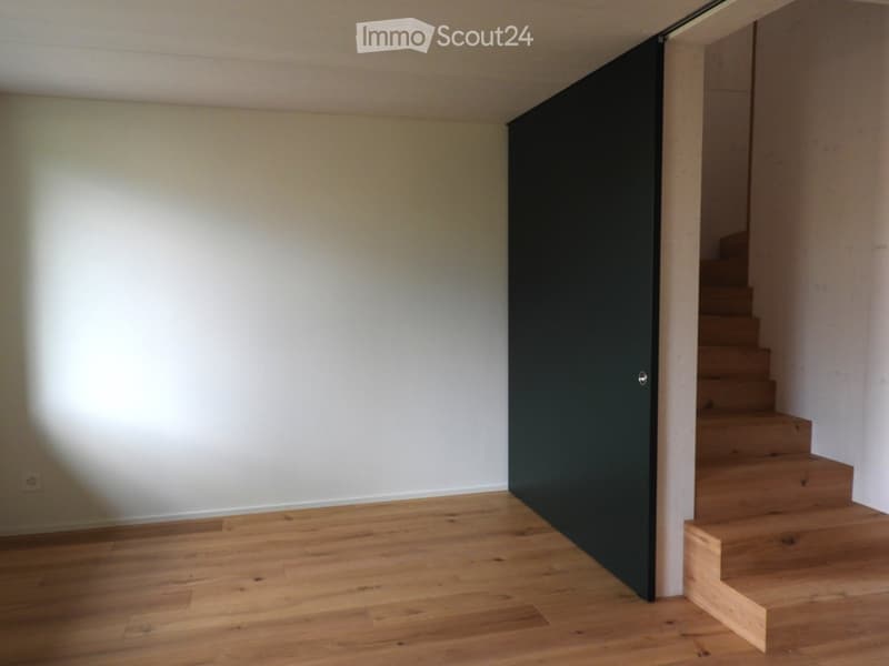 Empty hallway with light wooden flooring, white walls, and a black sliding door leading to a staircase.
