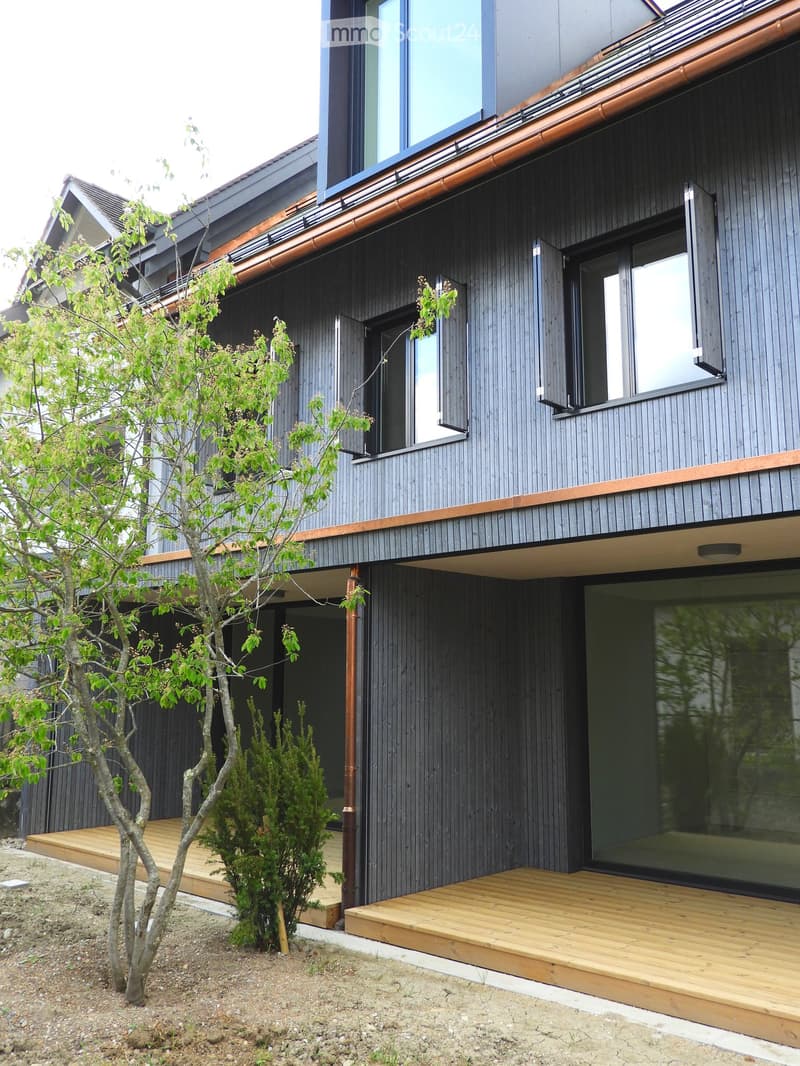 Modern two-story house with dark grey wooden siding, large windows, and a raised wooden deck area.