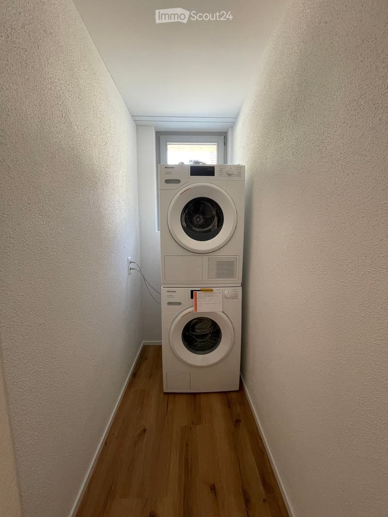 Narrow hallway with light wooden flooring and white walls, featuring a stacked washing machine and dryer unit against one wall.