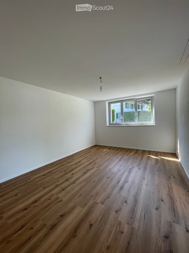 Empty room with wooden flooring and white walls, featuring a large window with a view of greenery outside.