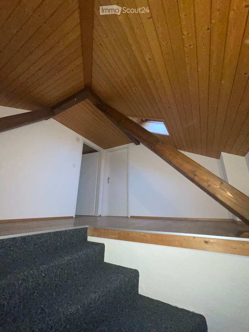 Staircase with wooden handrail and carpeted steps, leading to an open area with white walls and wooden ceiling beams.