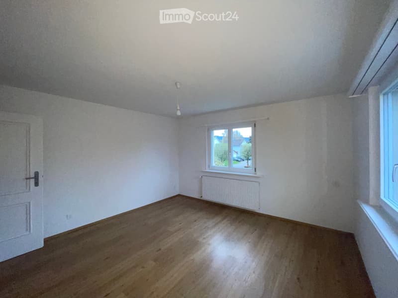 Empty room with wooden flooring, white walls, a single window with white frame, a radiator, and a door with a silver handle.