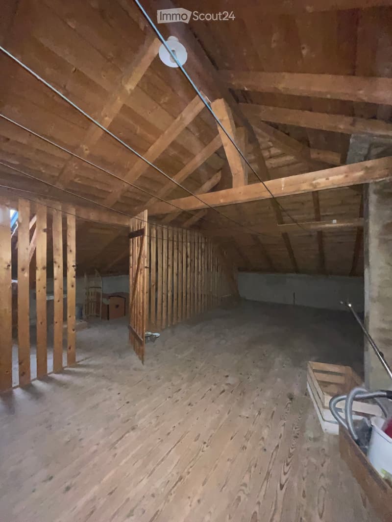 Empty attic space with exposed wooden beams and insulation, unfinished wooden floor, and a small open doorway leading to another area.