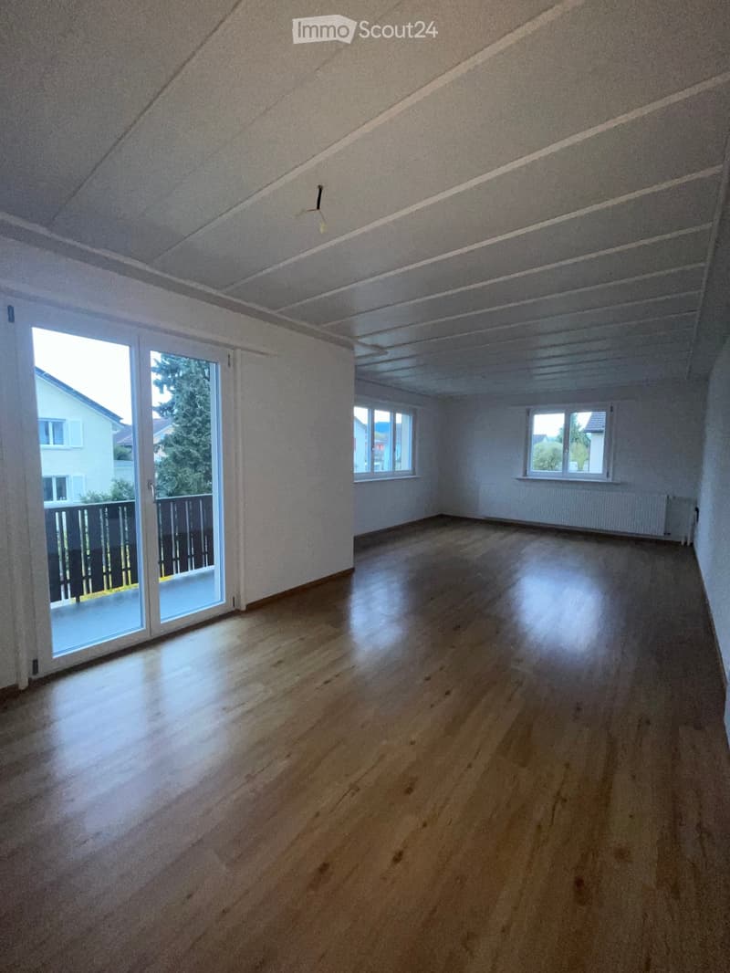 Empty room with wooden flooring, white walls, and large windows, including a sliding glass door leading to a balcony.