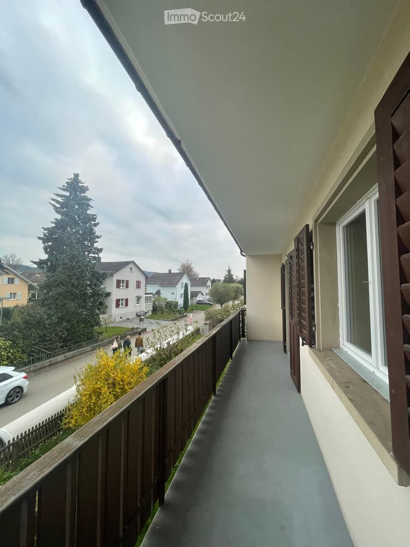 Balcony with wooden railing, grey flooring, and a view of a residential street with houses, trees, and parked cars.