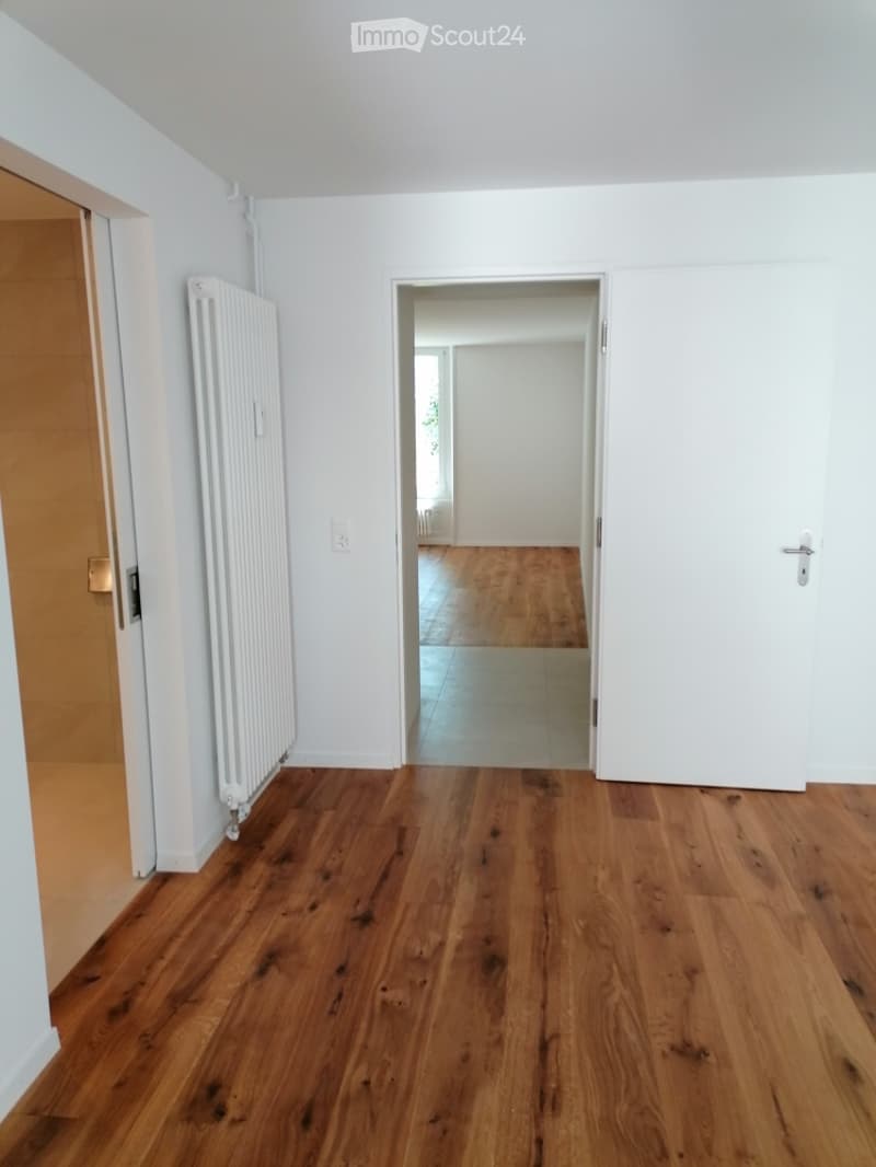 Empty hallway with wooden flooring, white walls, and a radiator on one side. A doorway leads to another room with similar flooring.