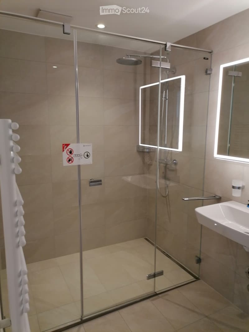 Modern bathroom with a glass-enclosed shower, a white sink, and a mirror with built-in lighting. Beige tiles and a towel rack are visible.